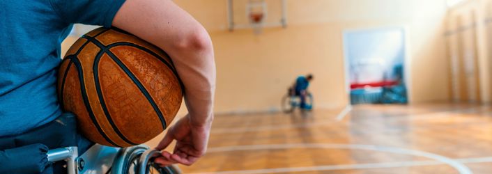 Photograph of person in a wheelchair holding a basketball. The background is out of focus but you can see a gymnasium with a net and another person in a wheelchair.