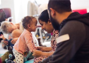 Photograph of baby girl standing up looking towards her mother who is holding her hands.