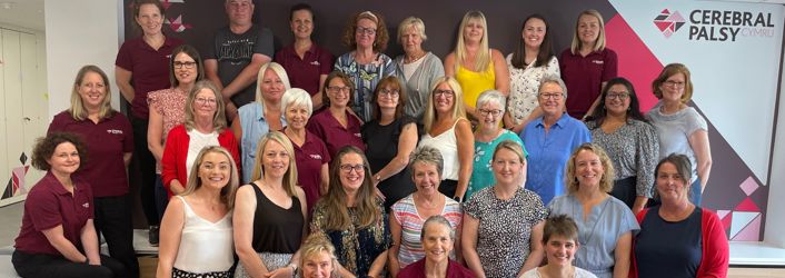 Photograph of all of our staff standing together in front of the Cerebral Palsy Cymru branded wall in our centre. 